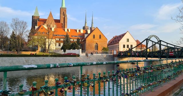 Brick churches with spires beside a river, a green iron bridge and railings covered in padlocks.